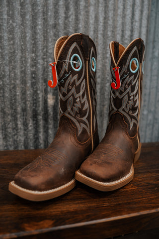 Pair of brown cowboy boots with decorative patterns on a wooden surface and metal wall background.