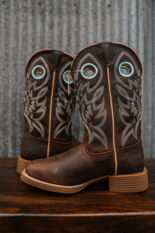 Pair of brown cowboy boots with decorative patterns on a wooden surface and metal wall background