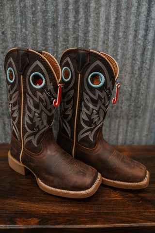 Pair of brown cowboy boots with decorative patterns on a wooden surface and metal wall background.
