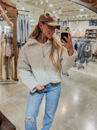Woman taking a mirror selfie wearing a light gray jacket, blue jeans, and a brown cap in a store.