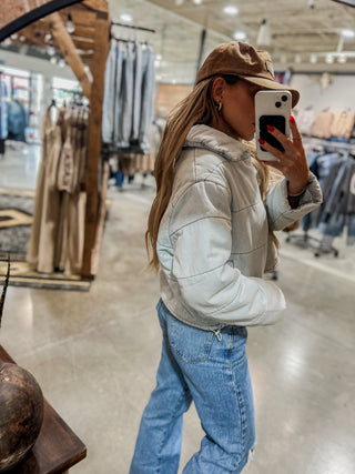 Person taking a mirror selfie wearing a light gray jacket, blue jeans, and a brown cap in a store.
