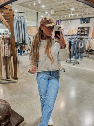 Woman taking a mirror selfie in a clothing store wearing a beige jacket, blue jeans, and a brown cap.