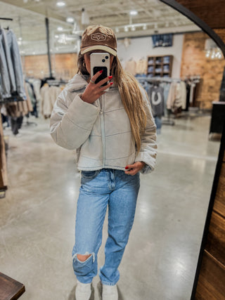 Person wearing a beige jacket, light blue jeans, and a cap in a store setting.