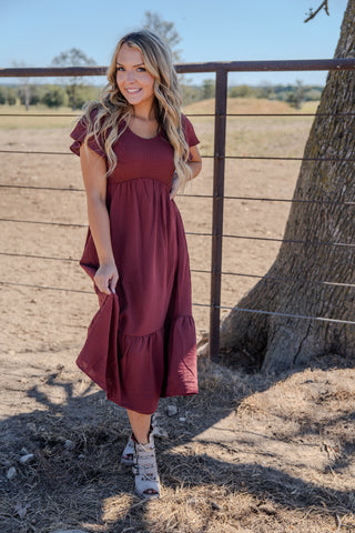 Woman in a burgundy dress standing outdoors near a wooden fence and tree.