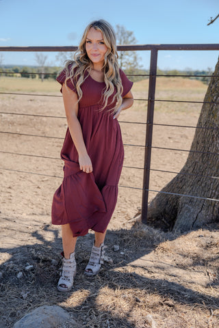 Woman in a burgundy dress standing near a wooden fence and tree in an outdoor setting.