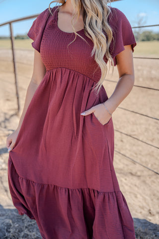 Woman wearing a burgundy dress standing outdoors with a fence and open field in the background.