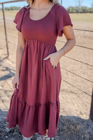 Woman wearing a maroon dress standing outdoors with a fence and field in the background