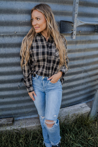 Woman wearing a plaid shirt and jeans standing against a corrugated metal wall.