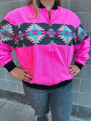 Person wearing a bright pink jacket with a geometric pattern, standing against a gray brick wall.