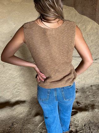 Person wearing a brown sleeveless top and blue jeans standing against a textured wall.