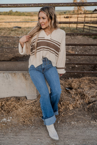 Woman sitting on a concrete block in a rural setting wearing a striped sweater and jeans.