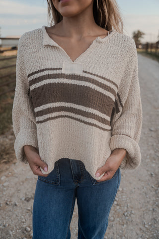 Person wearing a beige sweater with brown stripes and blue jeans on a gravel road.