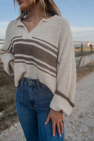 Person wearing a beige and brown striped sweater with blue jeans by a beach.