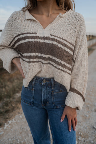 Person wearing a beige sweater with brown stripes and blue jeans outdoors.