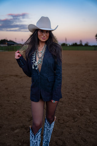 Woman in cowboy hat and boots standing in a dirt field with a sunset sky.