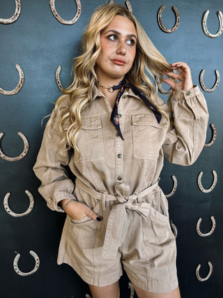 Woman in beige romper posing against a dark wall with horse shoe decorations