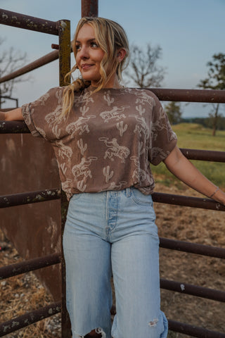 Woman wearing a patterned shirt and jeans standing behind a rusted metal gate.