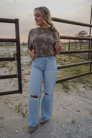 Woman standing in a rural setting wearing a patterned top and ripped jeans.