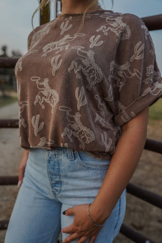 Person wearing a brown patterned top and light blue jeans standing near a fence.