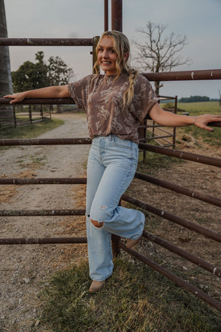Woman standing in a field with a metal gate, wearing a patterned top and light blue jeans.