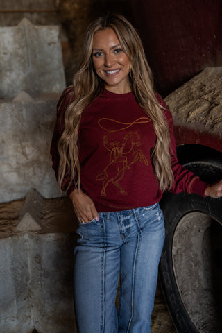 Woman wearing a maroon sweatshirt with a logo and blue jeans standing in front of rustic stone walls and a tire.