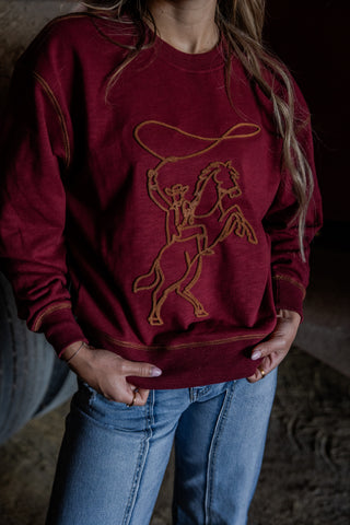 Person wearing a maroon sweatshirt with a horse and lasso design in a rustic setting