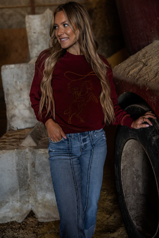 Woman wearing a maroon sweater with a graphic design and blue jeans, standing in a rustic setting.
