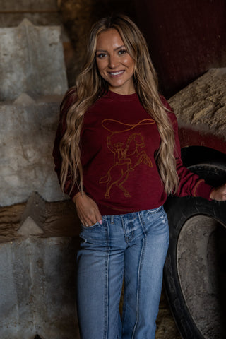 Woman wearing a red sweatshirt with a graphic design, standing in front of stone walls and tires.