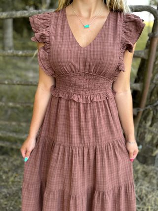 Woman wearing a brown checkered dress with ruffled sleeves in front of a rustic wooden fence.