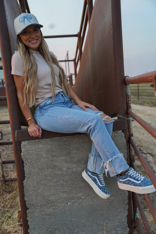 Woman sitting on a rusted metal structure wearing a cap, light shirt, jeans, and blue sneakers.
