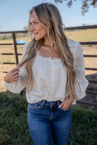 Woman in a white blouse and blue jeans standing outdoors with a fence and field in the background.