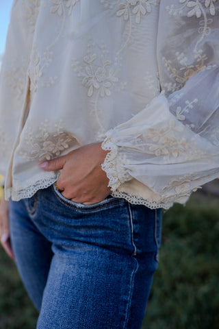 Person wearing a white lace blouse with long sleeves and blue jeans, standing outdoors.