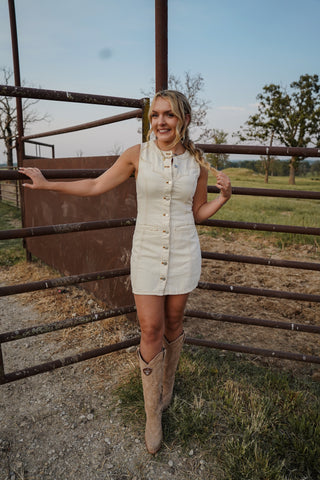 Woman in a white dress and cowboy boots standing next to a metal gate in an outdoor setting.