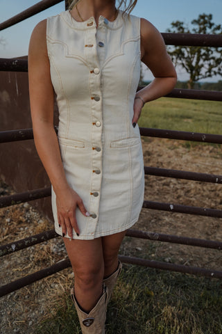 Person wearing a white denim dress with a button-up front, standing in a field with a metal fence.