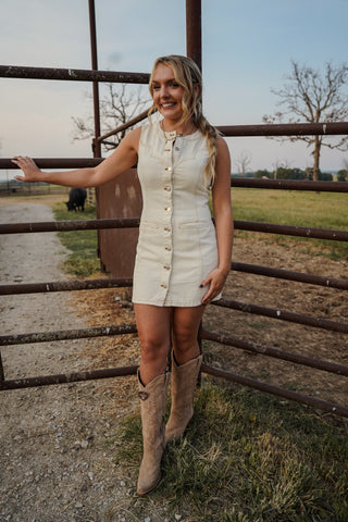 Woman in a white dress and beige boots standing near a wooden fence in an outdoor setting.
