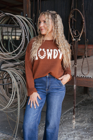 Woman wearing a brown 'HOWDY' sweater and blue jeans standing in front of rustic metal coils.