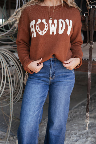 Person wearing a brown sweater with 'HOWDY' text and blue jeans in an indoor setting.