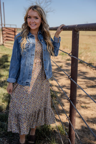 Woman in a floral dress and denim jacket standing next to a rustic fence in an outdoor setting.