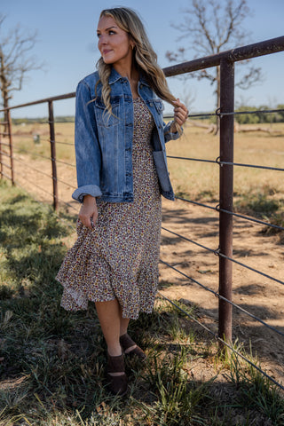 Woman in a floral dress and denim jacket standing near a fence in a rural setting