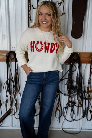 Woman wearing a white sweater with 'HOWDY' in red text, standing in front of horse equipment.