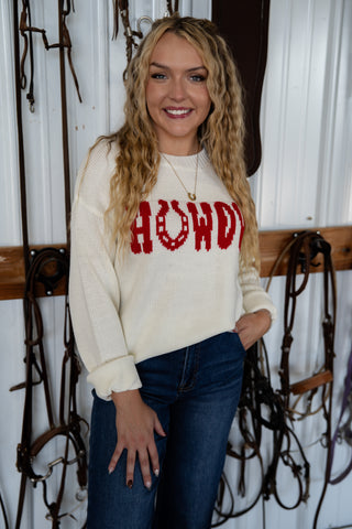 Woman wearing a cream sweater with 'HOWDY' in red text, standing in a barn setting.