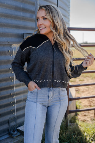 Woman wearing a black and gray zip-up sweater and light blue jeans standing against a metal wall.