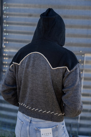 Person wearing a black and gray jacket with a hood, standing against a corrugated metal wall.