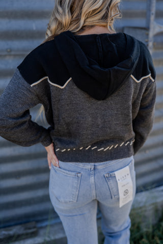 Person wearing a dark gray sweater with black hood and light blue jeans, standing against a corrugated metal wall.