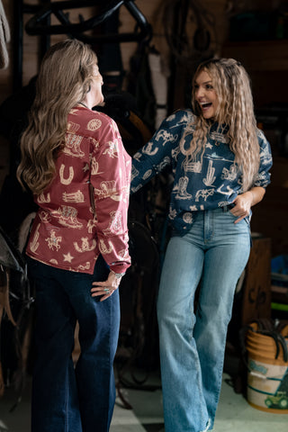 Two women standing in a rustic indoor setting, one wearing a red patterned shirt and blue jeans, the other in a blue patterned shirt and jeans.