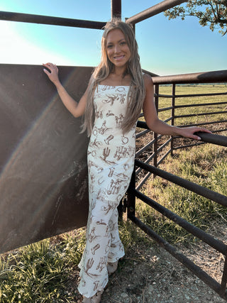 Woman in a white dress with patterns standing next to a metal fence in an open field.