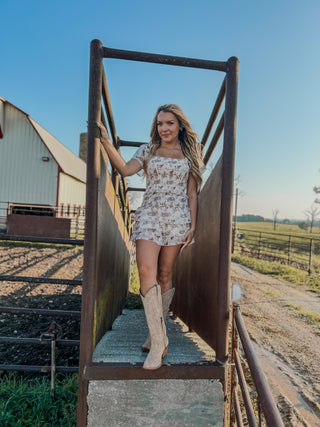 Woman in a floral dress and cowboy boots standing on a metal staircase in a rural setting.