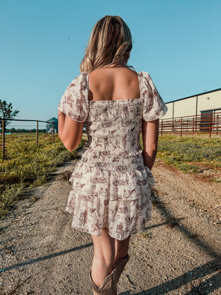 Woman in a floral dress walking on a dirt path with a clear blue sky and a building in the background.