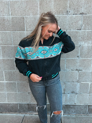 Woman wearing a black jacket with teal and white pattern standing against a brick wall.