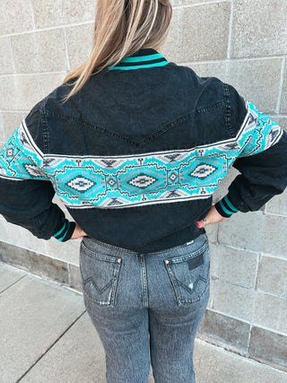 Person wearing a black jacket with a turquoise and white pattern, standing against a stone wall.